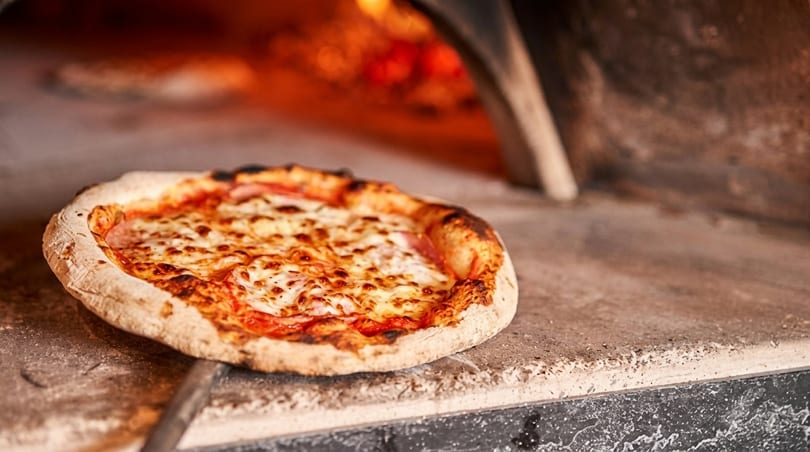 Freshly baked pizza with melted cheese and ham being removed from a wood-fired oven.