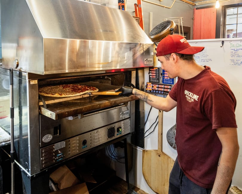 A Brickline Pizza Co. employee in a red hat and shirt slides a pizza into a large oven.